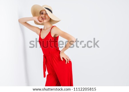 Beautiful girl in sun hat and red dress standing in studio on white background and looking at camera