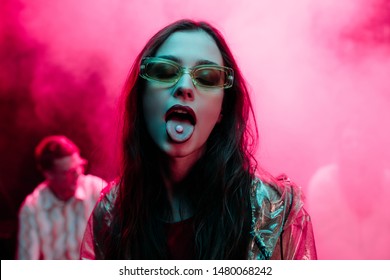 beautiful girl in sunglasses with lsd on tongue in nightclub with pink smoke