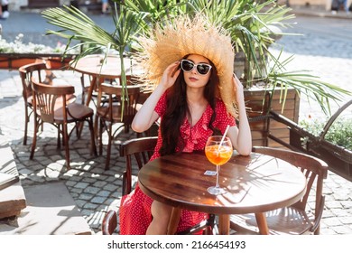 Beautiful Girl In Sunglasses, Straw Hat And Red Polka-dot Dress Sitting On The Summer Terrace With Aperol Spritz Drink. Young Woman Portrait