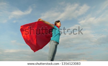 beautiful girl superhero standing on the field in a red cloak, cloak fluttering in the wind. Slow motion. girl dreams of becoming superhero. young girl standing in a red cloak expression of dreams