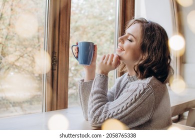 A beautiful girl in a sweater drinks tea or coffee against the background of a window and lights. Christmas or fall time