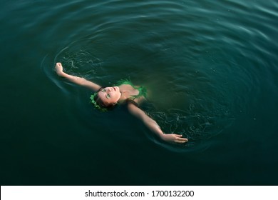 A Beautiful Girl In A Swimsuit Of Green Plants With A Wreath On Her Head Floats On Her Back In Troubled Water. Concept Art Of A Mistress Of The Lake From Legends.