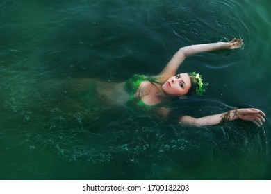 A Beautiful Girl In A Swimsuit Of Green Plants With A Wreath On Her Head Floats On Her Back In Troubled Water. Concept Art Of A Mistress Of The Lake From Legends.