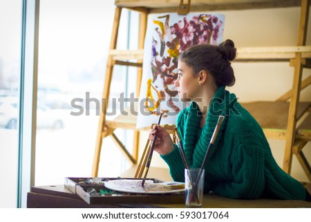 Beautiful girl thinks dreams with brushes at the easel, painting on canvas.