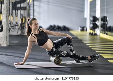 Beautiful girl is training with a black-yellow foam roller on the gray mat in the gym. She wears a black top with dark pants with prints and gray sneakers. Woman looks to the side. Horizontal.
