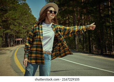 A beautiful girl traveler stands by a pine forest trying to catch a car on the road. Summer vacation outdoors. Tourism, hitchhiking.