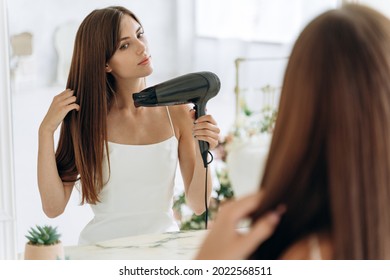 Beautiful girl using a hair dryer and smiling while looking at the mirror. Smiling woman drying hair with hair dry machine. Happy girl looking at mirror while using hair dryer in the bathroom 