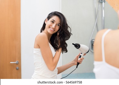 Beautiful girl using a hair dryer and smiling while looking at the mirror. Smiling woman drying hair with hair dry machine. Happy girl looking at mirror while using hair dryer in the bathroom.