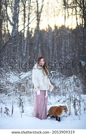 A beautiful girl walks with a fox in a snowy forest. Cold weather. Winter's Tale. Red fox. Photoshoot in a fairy-tale style.