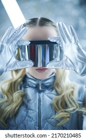 A Beautiful Girl With Wavy Blonde Hair Wearing A Silver Designer Jacket And Gloves Looks Through Her Trendy Futuristic Sunglasses. Silver Background With Neon Lamps. Fashion And Futuristic Style.