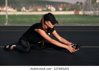 A Beautiful Girl Wearing Black Sportswear And A Black Cap Makes Warm Up Exercises At The Tennis Court Before Training.