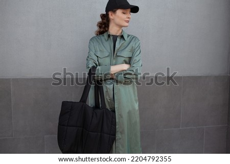 A beautiful girl is wearing a grey t-shirt, cloth cap, coat, black bag and jeans posing against the background of a wall. fashion outfit for the city, minimalism urban style clothing