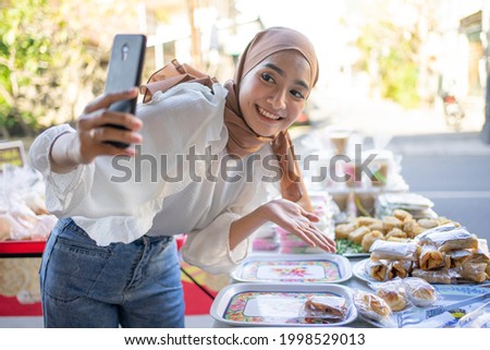 Beautiful girl wearing a smiling hijab using a mobile phone offers a variety of fried foods online at roadside stalls