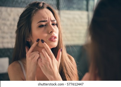 Beautiful girl in white bra is squeezing pimples on her face while looking into the mirror in bathroom