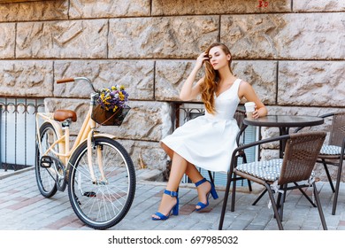 Beautiful Girl In White Dress Is Sitting At A Table Outside And Drinking Coffee On A Summer Day