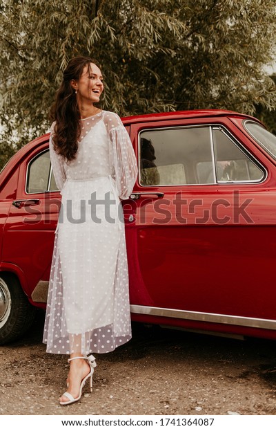 A\
beautiful girl in a white dress stands in the summer near a red\
retro car. Image with selective focus and\
toning