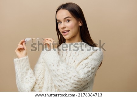 a beautiful girl in a white sweater poses on a light background, playfully twirling her glasses in her hands and looking at them from all sides, standing sideways and smiling happily with wide eyes