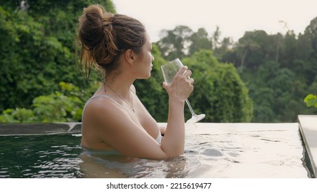 A Beautiful Girl In A White Swimsuit Standing In The Pool With A Wonderful View Takes A Glass Of Red Wine And Drinks And Looks Into The Distance With Pleasure. Film Grain Texture. Soft Focus.