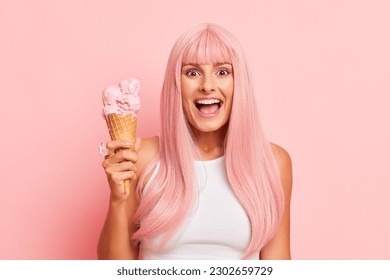 Beautiful girl in white top holds ice cream cornet in one hand, standing isolated on pink studio background, very happy face expression, copy space