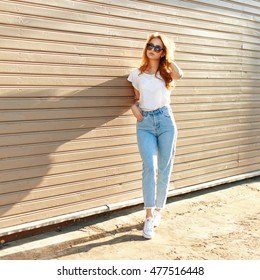 Beautiful girl in a white T-shirt, high waist jeans and white shoes standing near a wooden wall