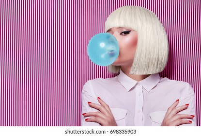 Beautiful girl in white wig blew up in pink gum bubble. A young girl in the studio on a background of a black and white vertical lines. Stylish girl wearing a white shirt.