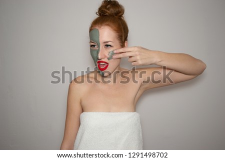 beautiful girl wrapped in a white towel with a clay mask on half of her face smears clay on the other half of her face and looks away on a gray white background, spa