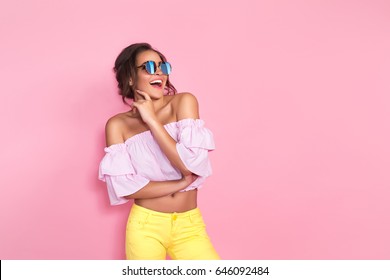 Beautiful girl in yellow jeans and pink shirt wearing sunglasses posing, screaming, smiling on pink background in studio. 