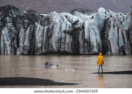 Beautiful girl in a yellow raincoat admires the powerful big Katla glacier during rainy weather in southern Iceland	