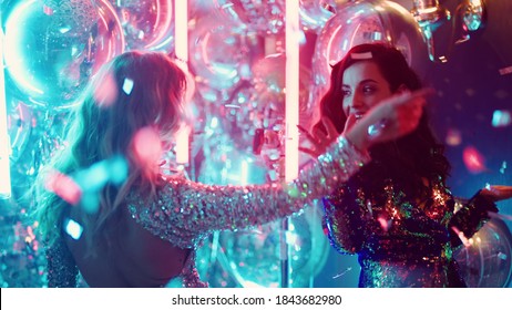 Beautiful girlfriends hugging in nightclub under confetti. Blonde and brunette girl friends spending night at party. Charming women dancing on neon lights background.