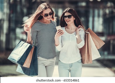 Beautiful girls in sun glasses are holding shopping bags, using a smart phone and smiling while standing outdoors