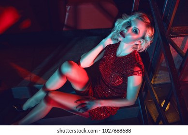 Beautiful Glam Blond Woman With Provocative Make Up Wearing Red Short Fitted Sequin Dress Sitting On The Stairs In The Night Club In Colourful Neon Lights. Text Space