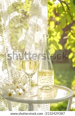 Beautiful glass and bottle of white wine with white flowers on glass table in summer garden in sunlight. Copy space. Summer drinks concept.