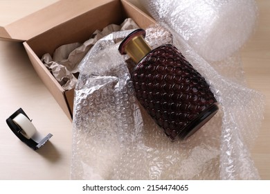 Beautiful glass vase in bubble wrap near cardboard box with paper and adhesive tape on wooden table
