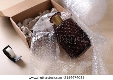 Beautiful glass vase in bubble wrap near cardboard box with paper and adhesive tape on wooden table