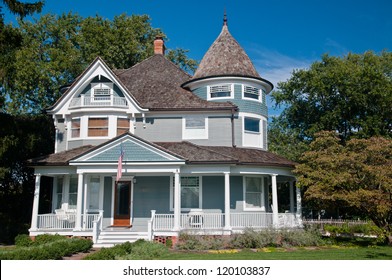Beautiful gray traditional victorian house.  American Flag hanging over the porch and shows a garden with flowers and trees.  Set against a cloudless blue sky