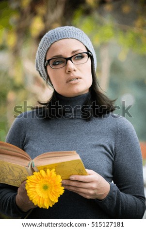 Beautiful green eyed young woman in warm clothes, vision glasses and gray hat smiling, reading a book and drinking tea outdoors in autumn time