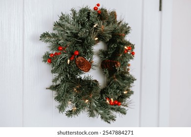 A beautiful green wreath featuring red berries and pinecones is elegantly displayed on a light-colored door, adding a festive touch to the entrance. Ideal for the holiday season. - Powered by Shutterstock