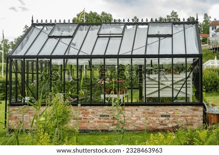 Beautiful greenhouse glass house in the garden yard near the villa. Lots of pots with different blooming plants. Greenhouse for growing plant seedlings. Landscape garden design.