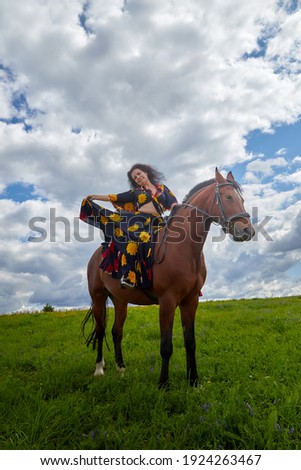 Beautiful gypsy girl on a horse in a field with green glass in summer day and blue sky and white clouds background. Model in ethnic dress posing with farm animal