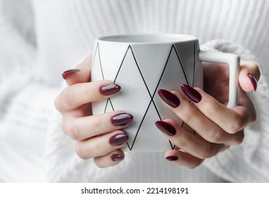 Beautiful hands of a young woman with dark red manicure on nails. Girl in a sweater holding a mug of tea