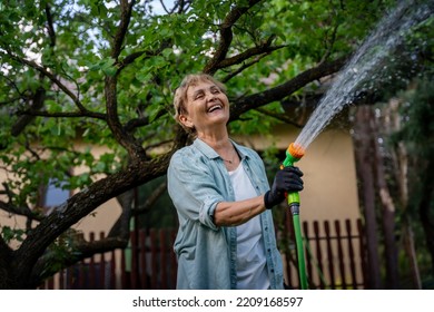 Beautiful Happy Active Elderly Woman Pensioner Watering Plants From A Hose In The Garden