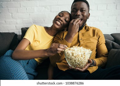 beautiful happy african american couple eating popcorn while sitting on sofa at home  