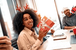 Beautiful Happy African American Excited Woman Mysteriously Holding A Christmas Present While Sitting At The Desk In The Office. Secret Santa, Exchanging Gifts Between Colleagues.