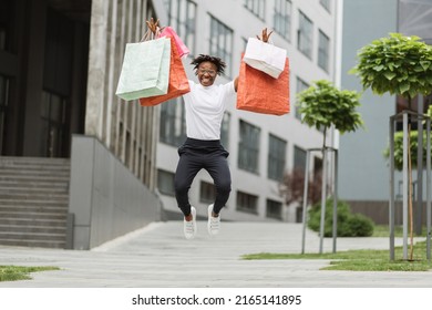 Beautiful Happy African Woman Wearing Black Pants And Stylish White T-shirt, Posing Outdoors In Beautiful Modern City With Many Colorful Shopping Bags While Jumping With Happiness New Purchases.