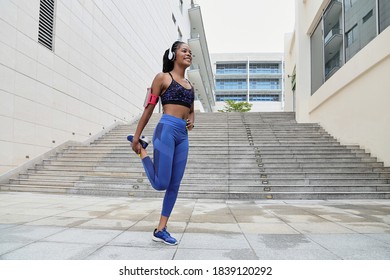 Beautiful happy African-American sportswoman in headphones standing on one leg and stretching quadriceps