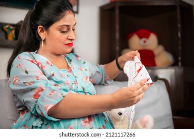 Beautiful Happy Asian, Indian Pregnant Woman Holding Newborn Baby Clothes In Hands And Looking At It With A Smile. She Is Sitting On Couch And Doing Preparation For The Baby’s Coming Out.