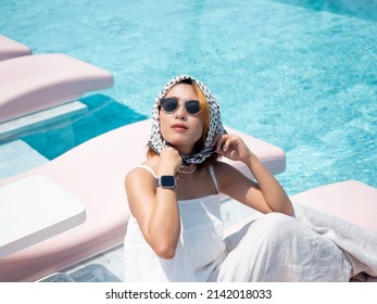 Beautiful Happy Asian Woman Portrait Look Cool Wearing Casual White Shirt, Sunglasses And Hair Scarf Closing Her Eyes While Sitting On Pink Sunbed Near The Pool On Sunny Day In Summer, Retro Style.