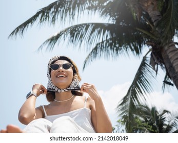 Beautiful Happy Asian Woman Portrait In Casual White Shirt Wearing Sunglasses And Hair Scarf, Enjoy With Smiling At The Beach Under The Coconut Palm Trees And Blue Sky Background In Summer.