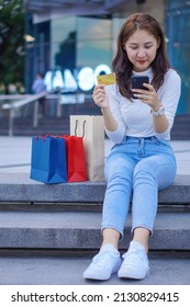 Beautiful happy Asian woman smiling cheerful and she holds a credit card and uses a smartphone to shop online with shopping bags on a shopping mall background.