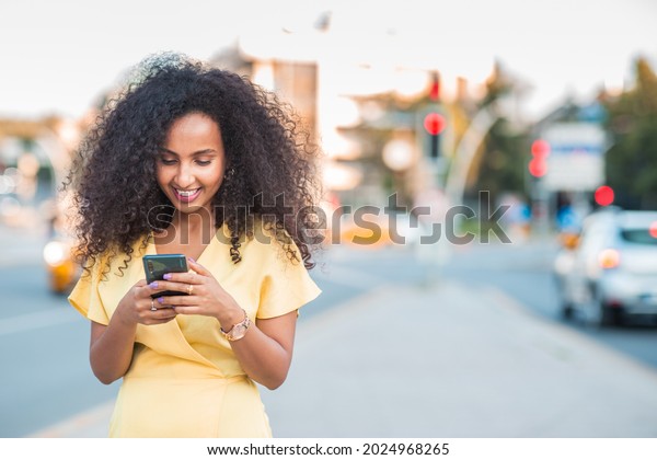 Beautiful happy Black Afro woman\
with positive and cheerful expression is looking her mobile phone\
in a city among cars and traffic lights, includes copy\
space.